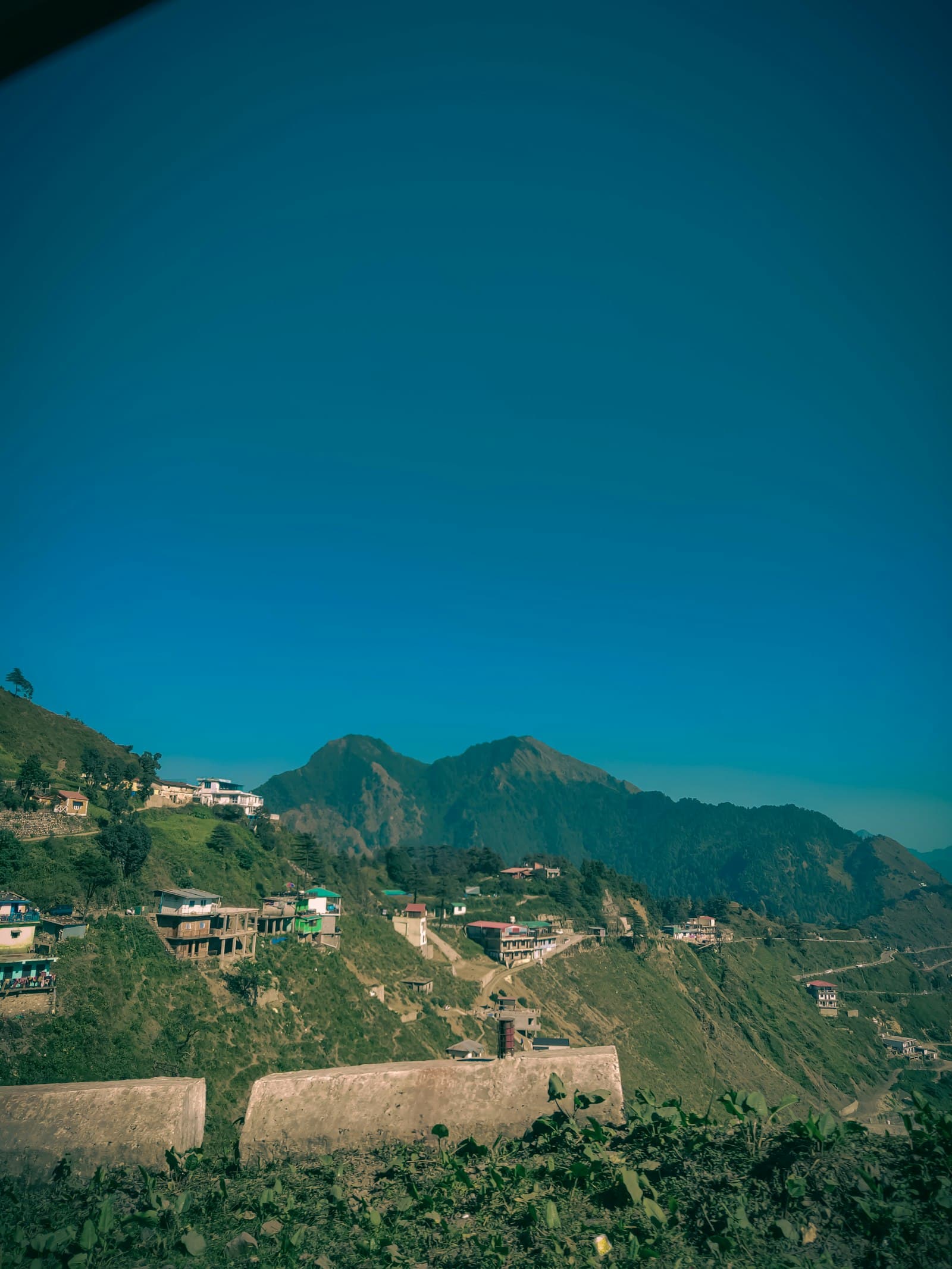 Houses scattered along a green hillside road below a forested peak under a clear blue sky